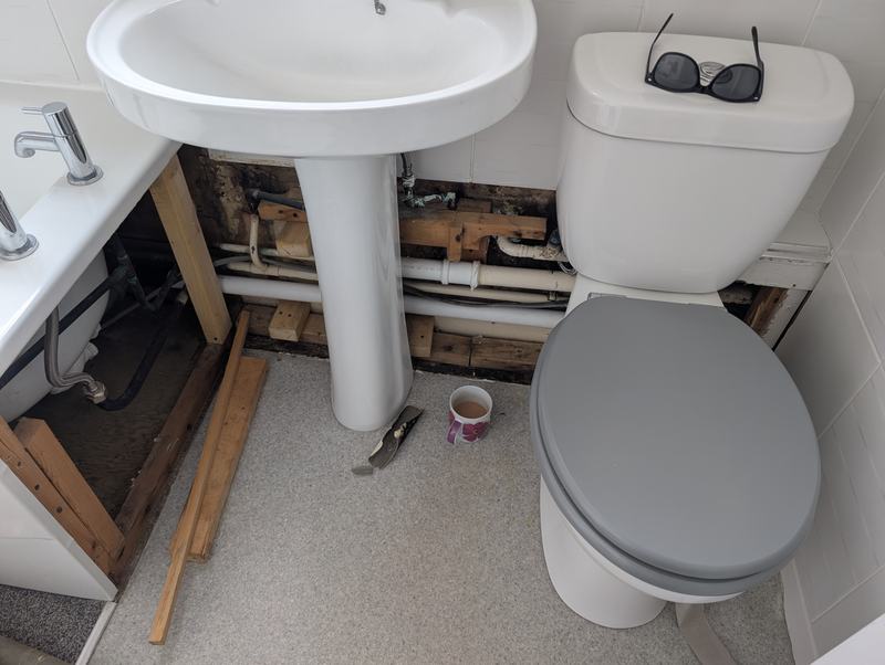 Exposed pipework and damaged timber behind a bathroom basin and toilet