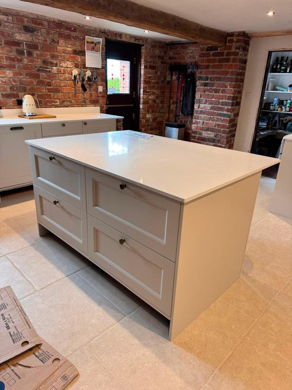 Kitchen island with four painted drawer units and a white worktop, in a room with exposed brick and an oak beam